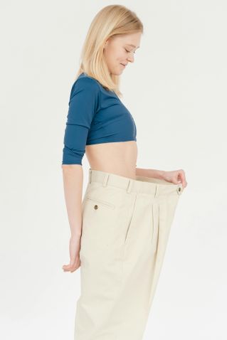 Side view of slim positive female with bare belly wearing oversized trousers standing on white background in light studio after weight loss